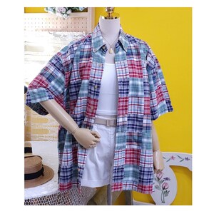 May include: A short-sleeved, patchwork plaid shirt in red, blue, green, and white. The shirt is buttoned and worn over a white vest top and shorts. A beige belt and a gold necklace complete the look. The shirt is displayed on a mannequin.