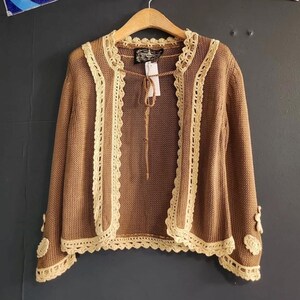 May include: Brown open-front cardigan with cream-colored lace trim. The long-sleeved sweater features a delicate crochet design along the edges and around the cuffs. A small tie closure is at the neckline. The cardigan is hanging on a wooden hanger.