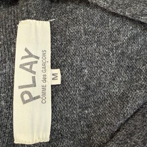 May include: Close-up of a gray knit garment with a white tag. The tag reads "PLAY COMME des GAR&Ccedil;ONS" with the size "M" indicated. The fabric appears soft and textured.