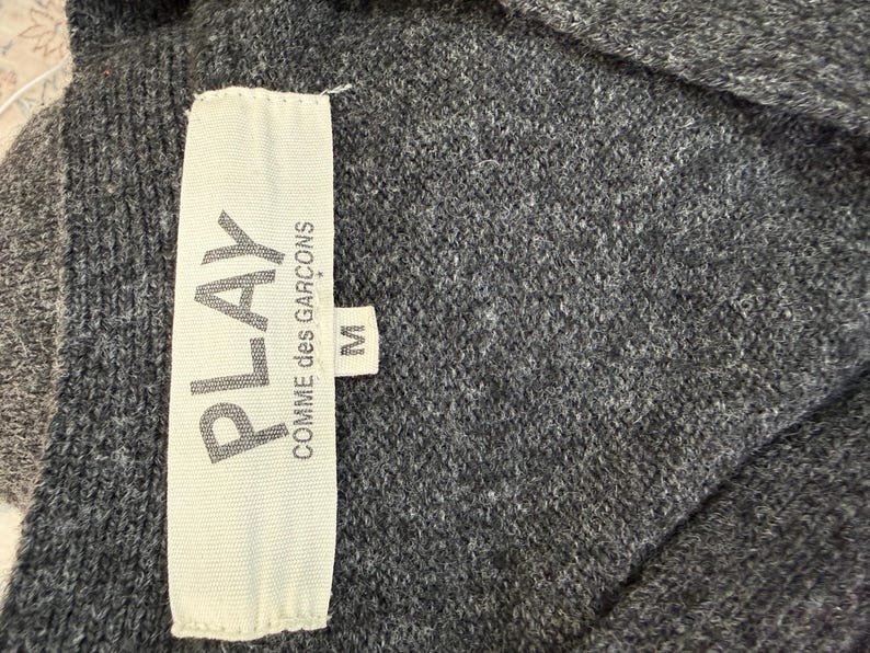 May include: Close-up of a gray knit garment with a white tag. The tag reads "PLAY COMME des GAR&Ccedil;ONS" with the size "M" indicated. The fabric appears soft and textured.