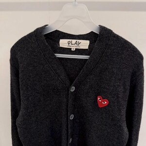 May include: Dark gray cardigan sweater with a button-up front. The sweater features a small red heart with eyes on the left chest. The label reads "PLAY COMME des GAR&Ccedil;ONS".