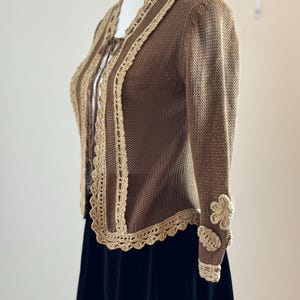 May include: A brown knitted cardigan with beige crochet trim and flower accents on the sleeves. The cardigan has a scalloped edge and a tie closure. The garment is displayed on a mannequin.