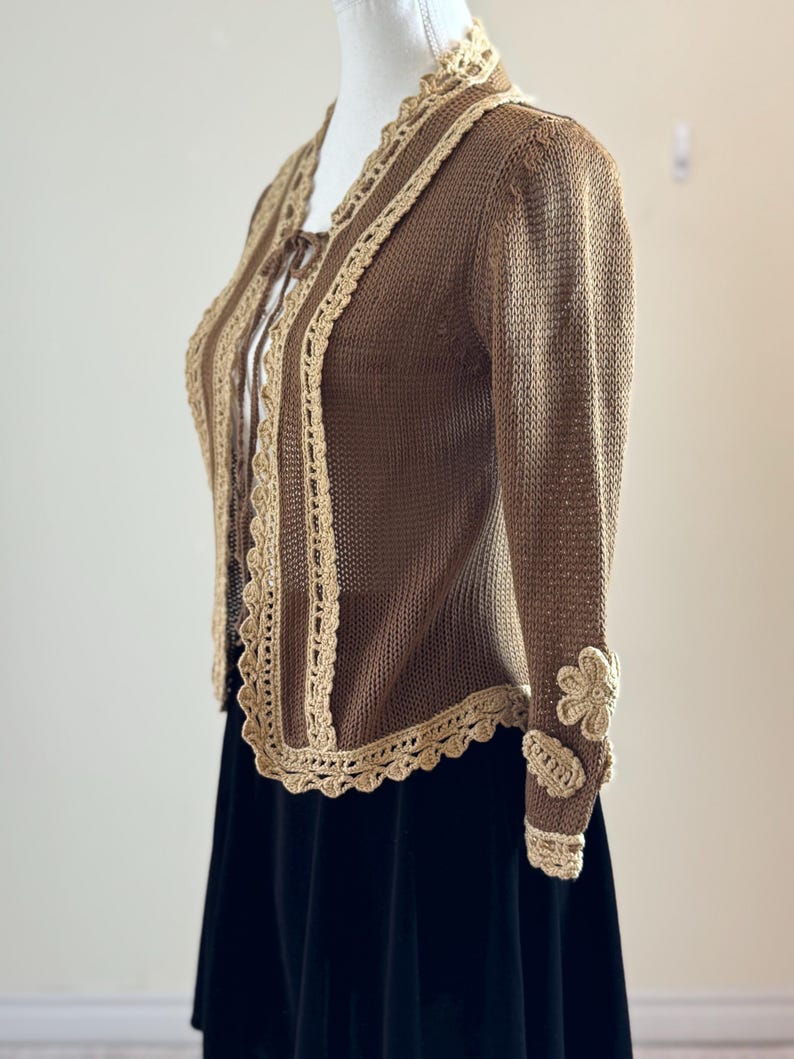 May include: A brown knitted cardigan with beige crochet trim and flower accents on the sleeves. The cardigan has a scalloped edge and a tie closure. The garment is displayed on a mannequin.