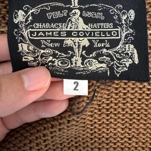 May include: A close-up of a brown knit fabric with a black label. The label reads "FELT LUSH, CHARACTER HATTERS, JAMES COVIELLO, New York" in white text. A white tag with the number "2" is attached.