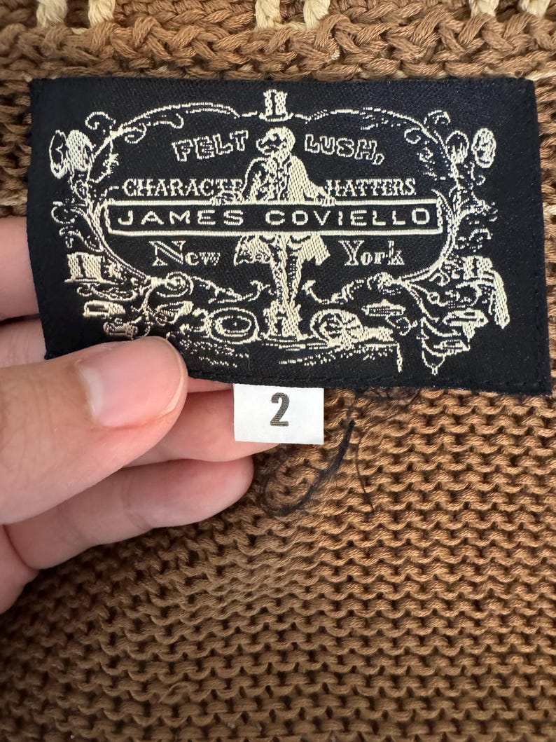 May include: A close-up of a brown knit fabric with a black label. The label reads "FELT LUSH, CHARACTER HATTERS, JAMES COVIELLO, New York" in white text. A white tag with the number "2" is attached.