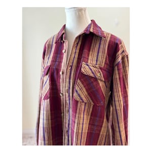 May include: A button-down shirt with a plaid pattern in burgundy, tan, and purple. The shirt has two chest pockets and a collared neckline. The fabric appears to be a soft, woven material.