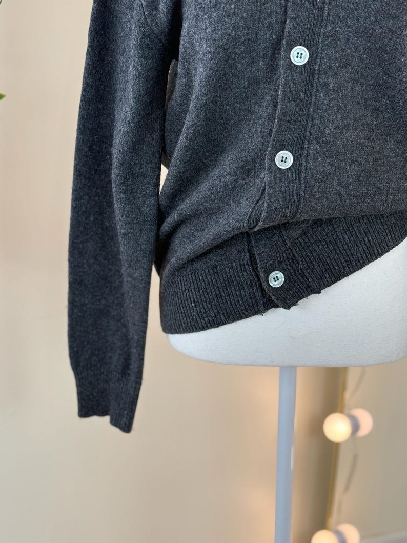 May include: A dark gray cardigan sweater with a button-up front. The sweater has a ribbed hem and cuffs, and the buttons are a light color. The fabric appears to be a soft knit material.