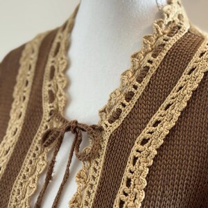 May include: A brown knit cardigan with tan crochet trim and a tie closure. The cardigan features vertical brown stripes with tan crochet lace detailing along the edges and neckline. The tie closure is made of brown yarn.