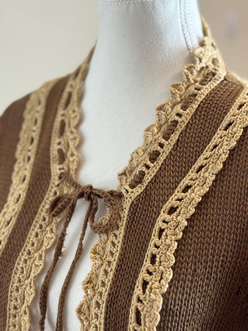 May include: A brown knit cardigan with tan crochet trim and a tie closure. The cardigan features vertical brown stripes with tan crochet lace detailing along the edges and neckline. The tie closure is made of brown yarn.
