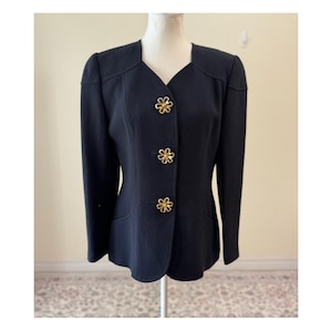 Könnte beinhalten: Ein schwarzer Blazer mit V-Ausschnitt und drei goldfarbenen Knöpfen in Blumenform. Der Blazer hat eine taillierte Passform und dezente Schulterpolster. Das Kleidungsstück wird auf einer Schaufensterpuppe präsentiert.
