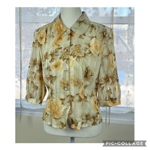 May include: A vintage floral blouse featuring a cream backdrop and a pattern of yellow and brown roses. The blouse has a collared neckline, pearl-like buttons, and three-quarter sleeves. The fabric has vertical stripes.