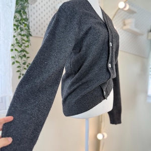 May include: A dark gray cardigan sweater with a V-neck and long sleeves. The sweater has a button-up front with white buttons. The garment is displayed on a white mannequin.