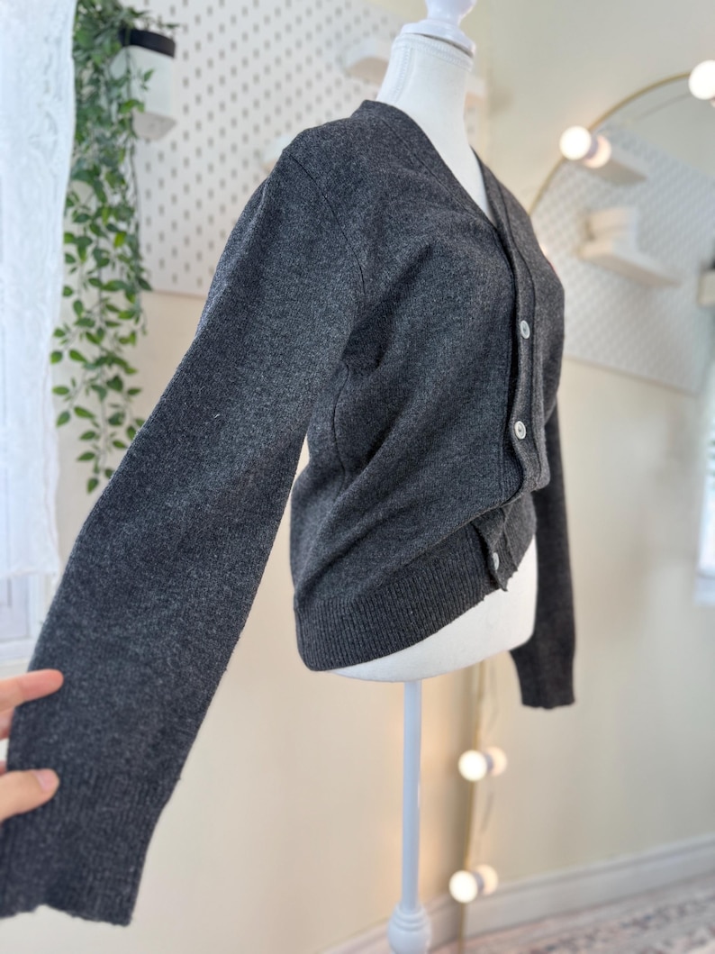 May include: A dark gray cardigan sweater with a V-neck and long sleeves. The sweater has a button-up front with white buttons. The garment is displayed on a white mannequin.