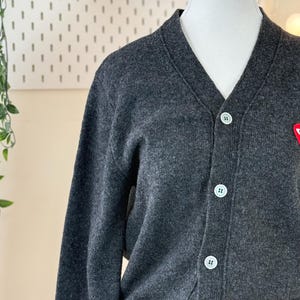 May include: A dark gray cardigan sweater with a V-neck and white buttons. The sweater has a small red emblem on the chest. The cardigan is displayed on a mannequin.