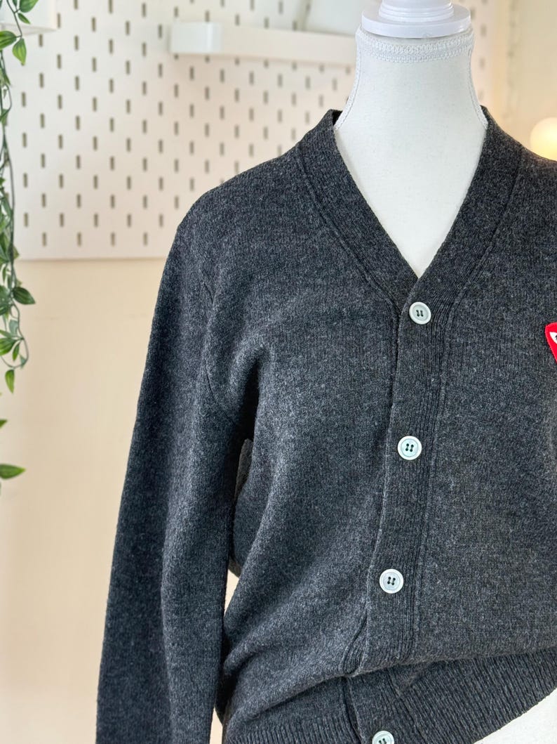 May include: A dark gray cardigan sweater with a V-neck and white buttons. The sweater has a small red emblem on the chest. The cardigan is displayed on a mannequin.