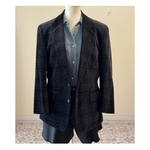 May include: A dark gray plaid blazer with a notched lapel and two front pockets. The blazer is worn over a light blue denim shirt and dark gray shorts. The blazer is displayed on a mannequin.