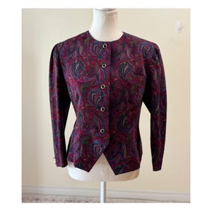 Puede incluir: Chaqueta vintage con estampado de cachemira en tonos burdeos, morado, verde y azul. La chaqueta presenta un cuello redondo, mangas largas y botones dorados en la parte delantera. La chaqueta se exhibe en un maniquí.