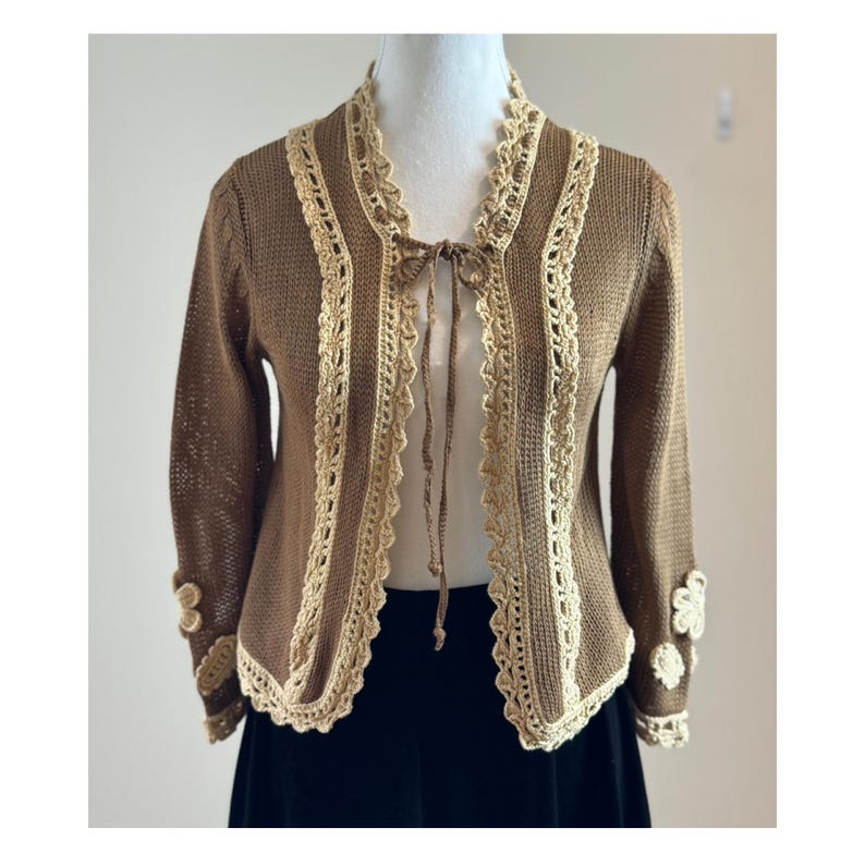 May include: A brown knitted cardigan with cream-colored crochet trim and a tie closure. The sleeves have decorative floral accents at the cuffs. The cardigan is displayed on a mannequin.