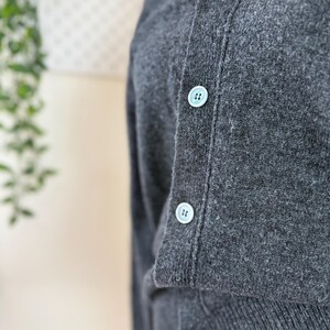 May include: Close-up of a charcoal gray cardigan sweater with light blue buttons. The sweater has a V-neck and is made of a soft, textured material. The buttons are evenly spaced down the front of the sweater.