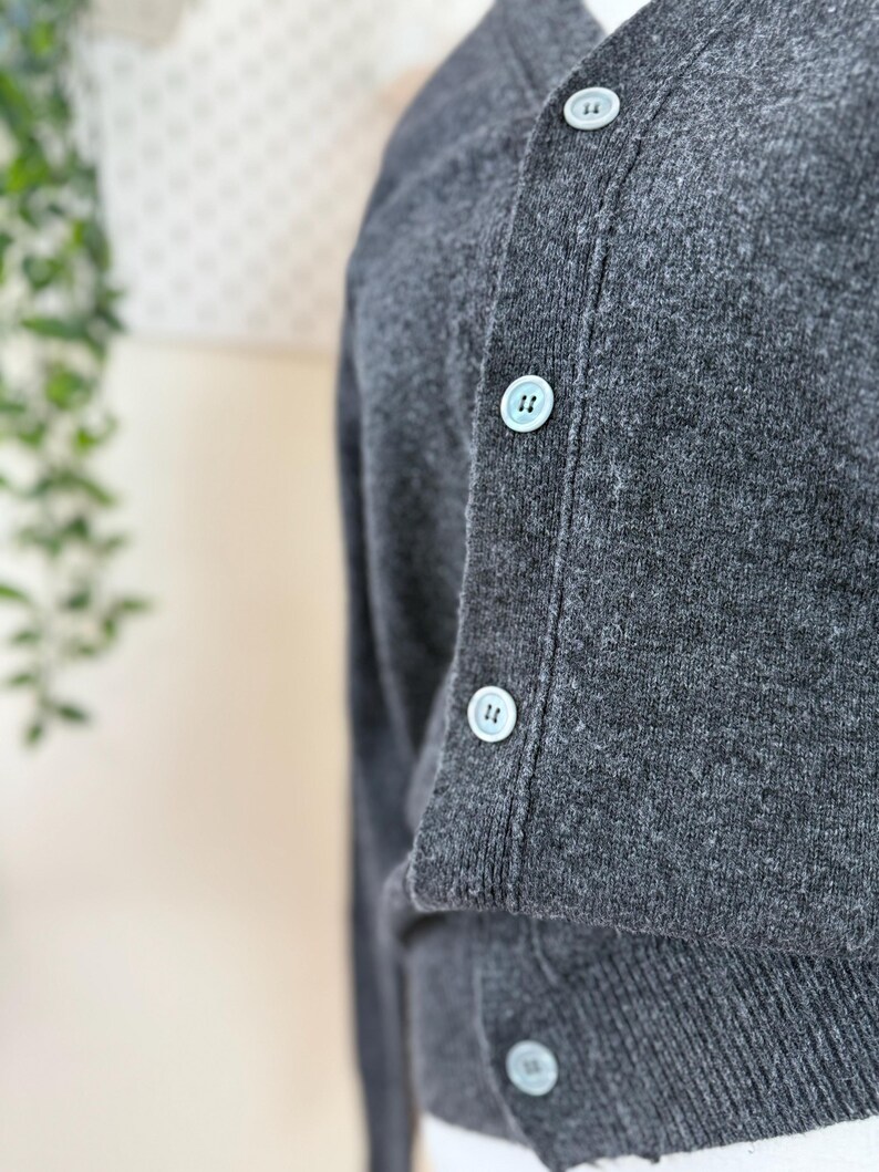 May include: Close-up of a charcoal gray cardigan sweater with light blue buttons. The sweater has a V-neck and is made of a soft, textured material. The buttons are evenly spaced down the front of the sweater.