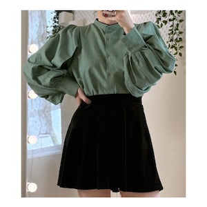 May include: A green striped blouse with long, puffed sleeves and a mandarin collar is paired with a black velvet mini skirt. The blouse is buttoned down the front. The skirt is pleated.