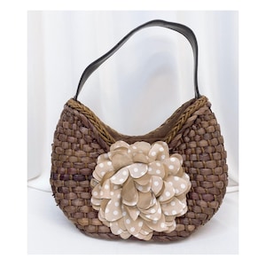May include: A brown woven handbag with a black handle and a large flower applique. The flower is beige with white polka dots. The bag has a curved shape and a braided trim.