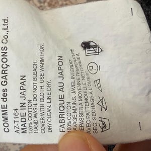 May include: A white tag with black text from Comme des Gar&ccedil;ons Co., Ltd. reads "MADE IN JAPAN" and "FABRIQUE AU JAPON". The tag includes washing instructions and fabric content.