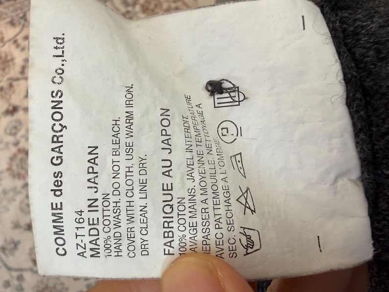 May include: A white tag with black text from Comme des Gar&ccedil;ons Co., Ltd. reads "MADE IN JAPAN" and "FABRIQUE AU JAPON". The tag includes washing instructions and fabric content.