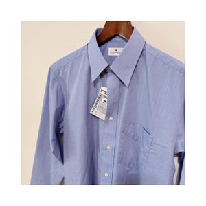Puede incluir: Una camisa azul claro con botones, bolsillo en el pecho y mangas largas. La camisa est&aacute; colgada en una percha de madera. Una etiqueta con el texto "MiracleCare" est&aacute; adjunta.