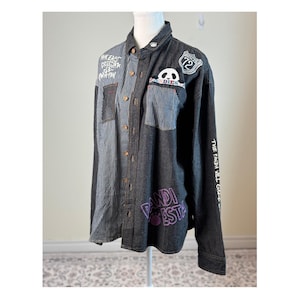 May include: A denim shirt with a patchwork design. It features vertical striped panels, solid denim, and patches with text and panda graphics. The sleeves have text, and the shirt has a collared neckline.