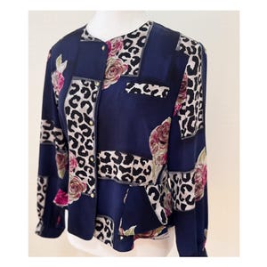 Puede incluir: Blusa azul marino con un patrón único. La blusa presenta una combinación de estampado de leopardo, diseños florales y formas geométricas. La parte superior de manga larga tiene botones dorados y se muestra en un maniquí.
