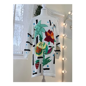 May include: White long-sleeve sweater dress with a colorful tropical fruit and flower design. The design includes a red flower, green palm leaves, and a strawberry. Black lines and blue dots accent the design.
