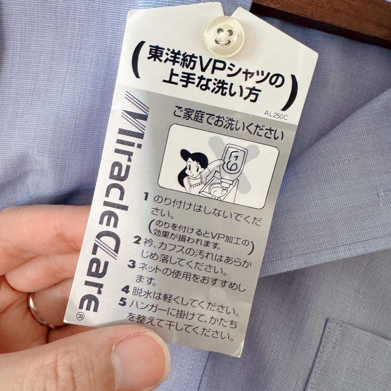 Puede incluir: Camisa azul claro con una etiqueta de cuidado adjunta. La etiqueta presenta texto japon&eacute;s y una ilustraci&oacute;n de una mujer lavando ropa. La etiqueta tambi&eacute;n tiene el texto "Miracle Care".