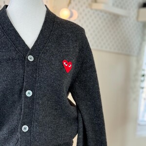 May include: Dark gray cardigan sweater with a V-neck and white buttons. A red heart-shaped patch with eyes is on the left chest. The sweater is displayed on a mannequin.