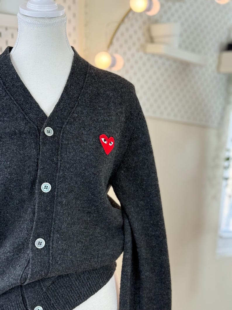 May include: Dark gray cardigan sweater with a V-neck and white buttons. A red heart-shaped patch with eyes is on the left chest. The sweater is displayed on a mannequin.