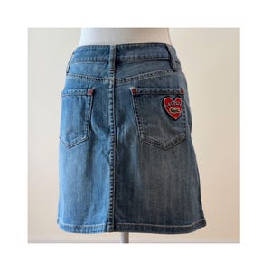 May include: Light blue denim skirt with a classic five-pocket design. Features a red heart-shaped patch with the Lacoste logo on the back pocket. The skirt is displayed on a mannequin.