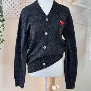 May include: A dark gray cardigan sweater with a V-neck and button closure. The sweater features a small red heart emblem on the left chest. The sweater is displayed on a mannequin.