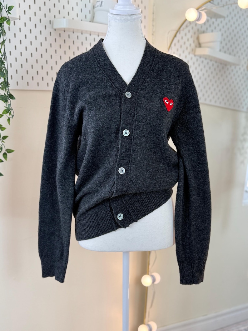 May include: A dark gray cardigan sweater with a V-neck and button closure. The sweater features a small red heart emblem on the left chest. The sweater is displayed on a mannequin.