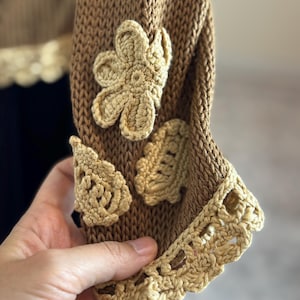 May include: Close-up of a brown knitted sleeve with beige crochet flower and leaf appliqu&eacute;s. The sleeve's cuff features a decorative beige crochet trim. The garment appears to be handmade, showcasing intricate details and textures.
