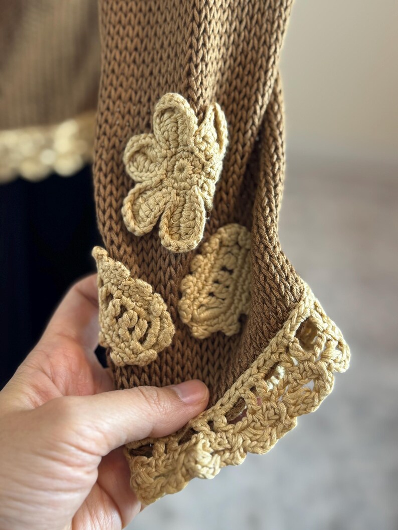 May include: Close-up of a brown knitted sleeve with beige crochet flower and leaf appliqu&eacute;s. The sleeve's cuff features a decorative beige crochet trim. The garment appears to be handmade, showcasing intricate details and textures.