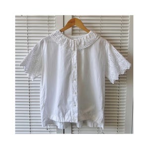 May include: A white short-sleeved blouse featuring an eyelet-detailed collar and sleeves. The blouse has a button-down front and a drawstring hem. The garment is displayed on a wooden hanger against a white shutter background.