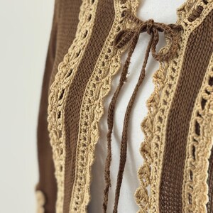 May include: A brown and beige crochet cardigan with a tie closure. The open-front sweater features vertical stripes of brown knit fabric and beige crochet lace trim. The sleeves are long and the cardigan is made of a soft, textured material.