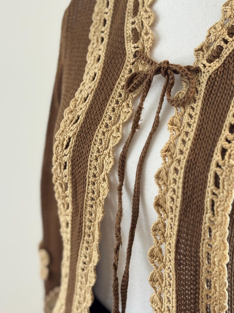 May include: A brown and beige crochet cardigan with a tie closure. The open-front sweater features vertical stripes of brown knit fabric and beige crochet lace trim. The sleeves are long and the cardigan is made of a soft, textured material.