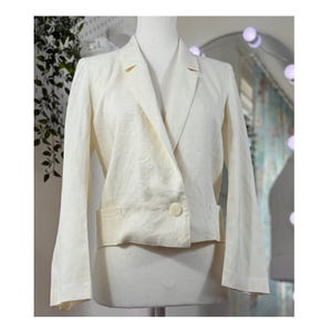 Könnte beinhalten: Ein cremefarbener Blazer mit einem Ein-Knopf-Verschluss und einem Revers. Der Blazer hat lange Ärmel und zwei Vordertaschen. Die Jacke wird auf einer Schaufensterpuppe präsentiert. Der Blazer ist aus leichtem Stoff.