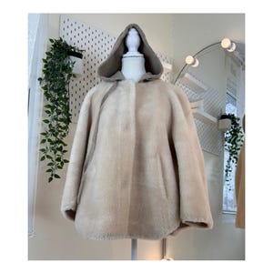 May include: A beige hooded cape coat displayed on a mannequin. The coat features a soft, textured exterior with a contrasting taupe-colored hood. The design includes a single button closure and a relaxed, flowing silhouette, suitable for outerwear.