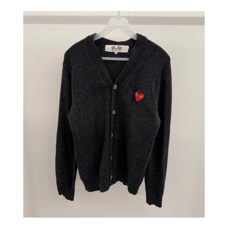 May include: Dark gray cardigan sweater with a V-neck and button closure. Features a red heart logo with eyes on the left chest. The label on the collar reads "PLAY".