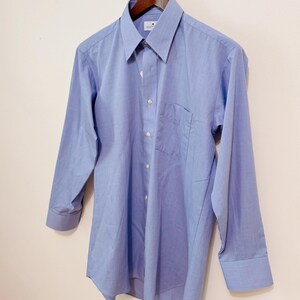 Puede incluir: Una camisa azul claro con botones, mangas largas, un bolsillo en el pecho y un cuello puntiagudo. La camisa est&aacute; colgada de una percha de madera. La camisa tiene botones blancos y un peque&ntilde;o logotipo en el cuello.