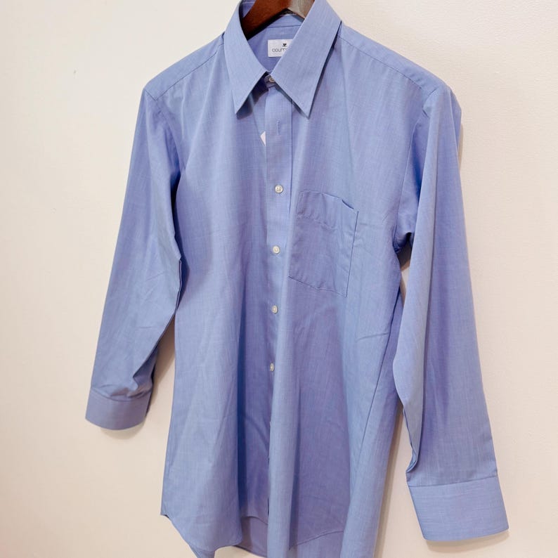 Puede incluir: Una camisa azul claro con botones, mangas largas, un bolsillo en el pecho y un cuello puntiagudo. La camisa est&aacute; colgada de una percha de madera. La camisa tiene botones blancos y un peque&ntilde;o logotipo en el cuello.