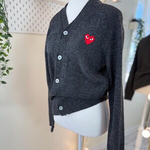 May include: Dark gray cardigan with a V-neck and button closure. Features a red heart-shaped patch with eyes on the left chest. The cardigan is displayed on a mannequin. The buttons are white.