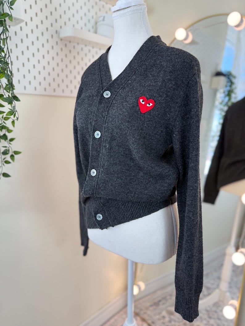 May include: Dark gray cardigan with a V-neck and button closure. Features a red heart-shaped patch with eyes on the left chest. The cardigan is displayed on a mannequin. The buttons are white.